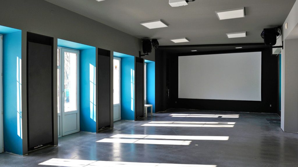 Zdjęcie wnętrza nowoczesnej, pustej sali konferencyjnej lub eventowej. Przestrzeń ma prostokątny kształt i jest utrzymana w industrialno-nowoczesnej estetyce. Podłoga wykonana jest z szarego, polerowanego betonu. Ściany boczne pomalowane są na niebieski kolor, z ciemnoszarymi pionowymi panelami akustycznymi lub dekoracyjnymi. Po lewej stronie sali znajdują się trzy wysokie, białe okna i drzwi, przez które wpada mocne dzienne światło, rzucając wyraźne, geometryczne smugi na podłogę. Ściana frontalna jest w całości czarna, matowa — na jej tle zamontowany jest duży, biały ekran projekcyjny. Po obu stronach ekranu widoczne są czarne głośniki ścienne. Pod sufitem zainstalowany jest projektor multimedialny. Sufit jasnoszary, oświetlony panelami LED wpuszczanymi w sufit. Sala jest pusta — bez mebli, z wyjątkiem jednego małego białego taboretu przy ścianie. Całość sprawia wrażenie przestrzeni przygotowanej do aranżacji pod różnego rodzaju wydarzenia.