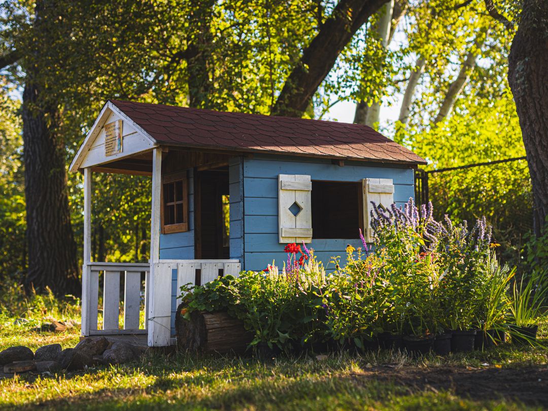 Drewniany domek ogrodowy dla dzieci wśród drzew, obok rabata z kolorowymi kwiatami i roślinami.