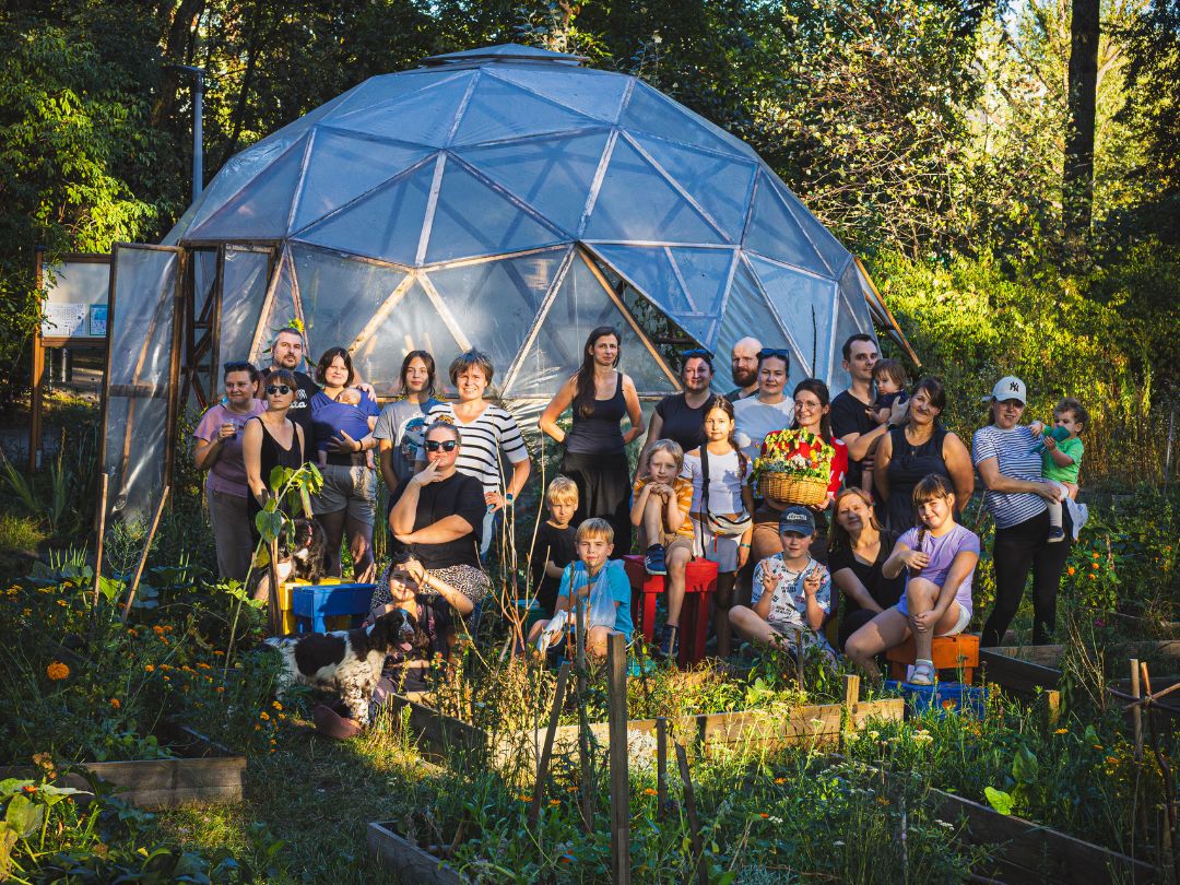 Grupa ogrodników pozująca przed szklarnią kopułową w ogrodzie społecznym, otoczona grządkami z warzywami i roślinami.