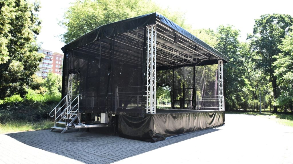 Mobilna scena plenerowa Domu Kultury „Świt" ustawiona w parku — scena estradowa na przyczepie z podwyższonym podestem obitym czarną tkaniną. Aluminiowa konstrukcja kratownicowa po bokach i nad sceną podtrzymuje zadaszenie kryte czarną plandeką. Na belkach sufitowych zamontowane reflektory. W głębi sceny przezroczyste panele tylnej ściany (backdrop), przez które widoczne są zielone drzewa. Na środku podestu metalowa barierka ochronna. W tle zielony park w słoneczny dzień.