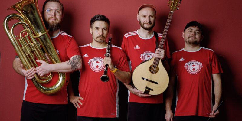 Na zdjęciu stoją czterej mężczyźni ustawieni w rzędzie na tle jednolitej, czerwonej ściany. Wszyscy mają na sobie czerwone koszulki sportowe i czarne spodenki. Każdy z nich trzyma instrument muzyczny: pierwszy z lewej dużą tubę, drugi klarnet, trzeci mandolinę, a czwarty pałki perkusyjne.