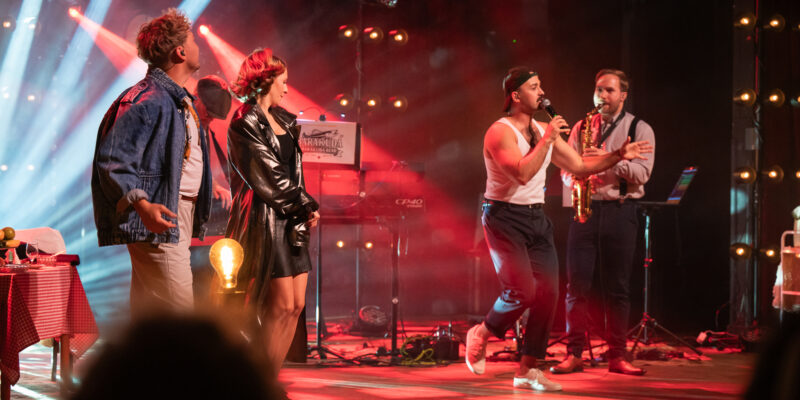 Kolorowe zdjęcie z występu na scenie. Czworo artystów stoi na oświetlonej czerwonymi i niebieskimi światłami scenie. Po lewej stronie mężczyzna w jeansowej kurtce i kobieta z rudymi włosami w czarnej skórzanej kurtce stoją zwróceni w stronę pozostałych wykonawców. Po prawej mężczyzna w białej koszulce bez rękawów śpiewa do mikrofonu w dynamicznej pozie, obok niego stoi saksofonista w kamizelce. W tle widoczna jest tablica z napisem „Barakuda" oraz sprzęt muzyczny. Na pierwszym planie głowy publiczności.