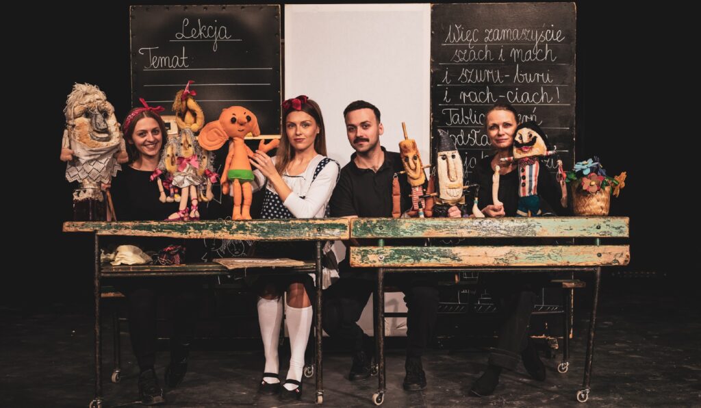 Cztery postaci siedzą w ławkach szkolnych. Na stole znajdują się lalki, a obok nich czarna tablica, tworząc scenę do zabawy lub edukacji.