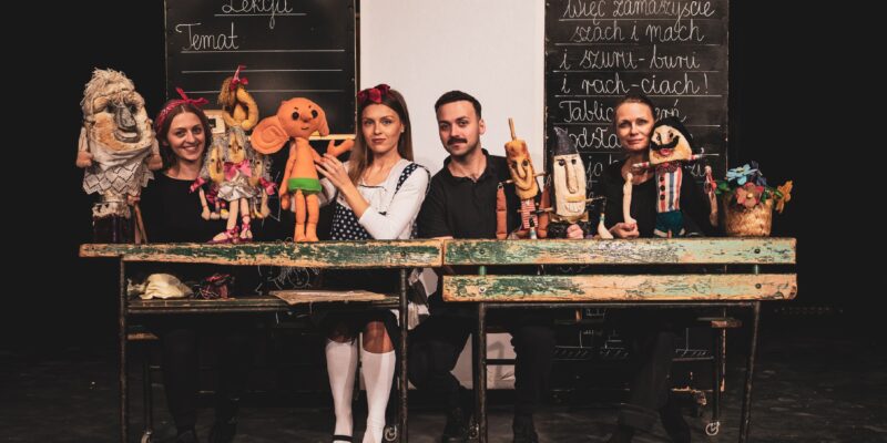 Cztery postaci siedzą w ławkach szkolnych. Na stole znajdują się lalki, a obok nich czarna tablica, tworząc scenę do zabawy lub edukacji.