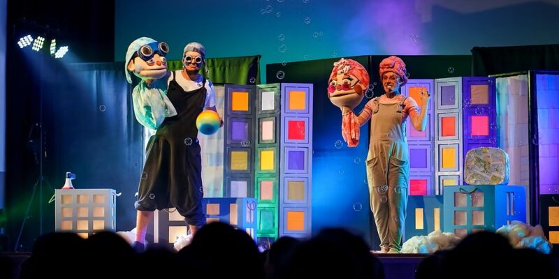 Na oświetlonej scenie teatralnej stoją dwie postacie w kostiumach i dużych maskach przedstawiających starsze osoby. W tle znajduje się kolorowa scenografia przypominająca budynki miasta, a wokół unoszą się bańki mydlane. Na dole widoczna jest publiczność oglądająca przedstawienie.
