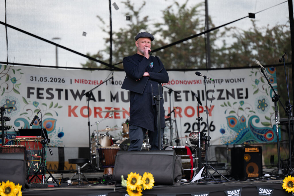 napisz tekst alternatywny do zdjęcia13:44Claude responded: Prowadzący na scenie Festiwalu Muzyki Etnicznej "Korzenie Europy 2026" w Parku Bródnowskim, w tle baner z datą 31.Prowadzący na scenie Festiwalu Muzyki Etnicznej "Korzenie Europy 2026" w Parku Bródnowskim, w tle baner z datą 31.05.2026 i nazwą festiwalu, na pierwszym planie żółte słoneczniki, w głębi scena z instrumentami perkusyjnymi.