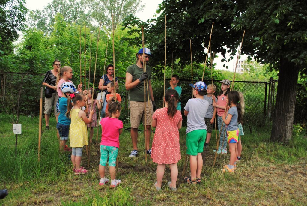 Wolontariusz i grupa dzieci stoją w kole i budują fasolowe tipi. W rękach trzymają bambusowe tyczki.