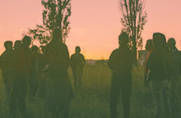 Grupa osób stojąca w kręgu na tle zachodzącego słońca.