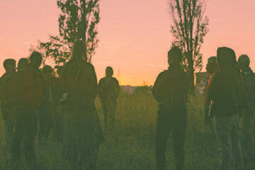 Grupa osób stojąca w kręgu na tle zachodzącego słońca.