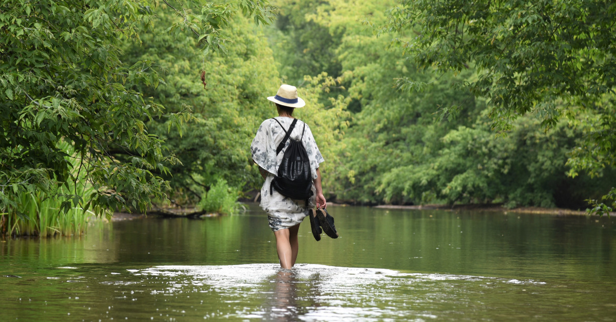 Kobieta w kapeluszu spaceruje rzeką Świder. W ręku trzyma buty.